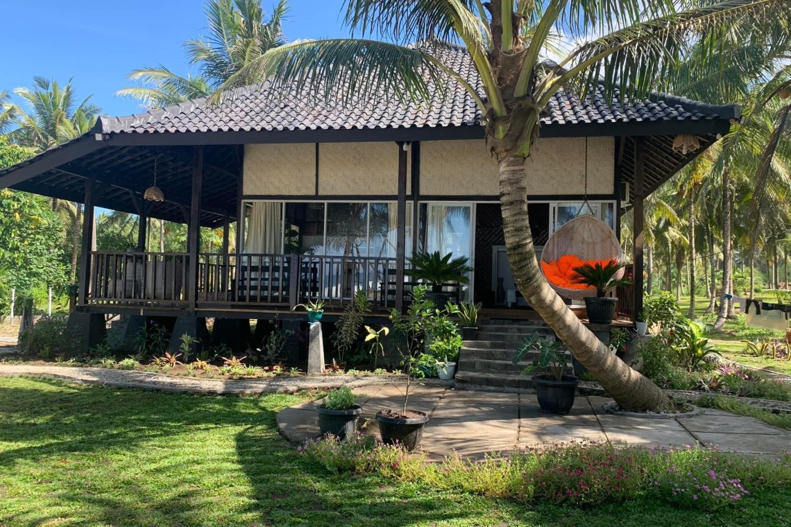 Traditional Balinese beachfront villa with black-trimmed veranda, signature orange egg chair, and lush palm grove surroundings